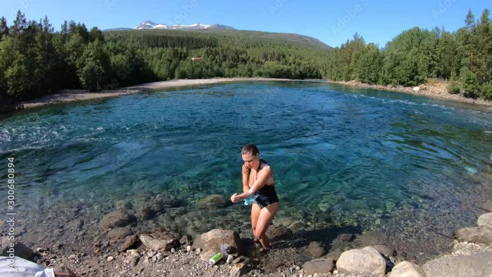 A young, attractive, brunette woman in a swimsuit is washing herself with a loofah and soap in a blue and turquoise colored river in the wild outdoors in Norway. A young, attractive, brunette woman in a swimsuit is washing herself with a loofah and soap in a blue and turquoise colored river in the wild outdoors in Norway.