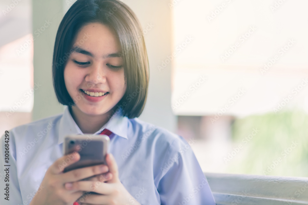 Smiling female Asian high school student in white uniform is enjoying social media on her smartphone. Smiling female Asian high school student in white uniform is enjoying social media on her smartphone.
