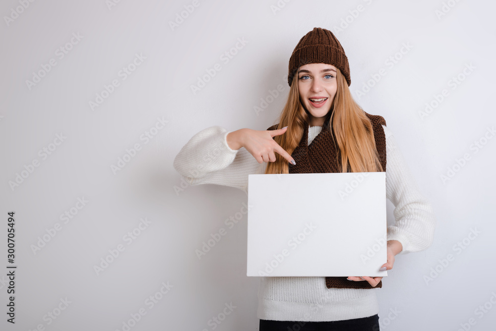 Naklejka premium Woman in winter clothes holds a white poster and points her finger on a gray background