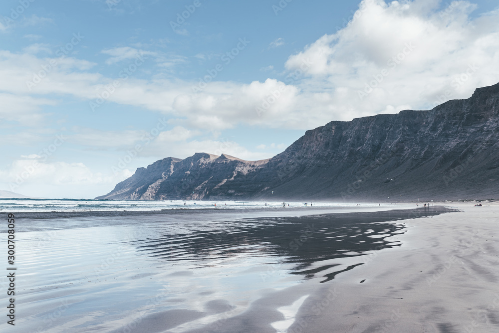 reflections in wet sand and Playa de Famara beach on Lanzarote, Canary Islands, against mountains, ocean and blue sky