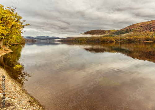 The mist beginning to clear as it drifts over Coniston lake in the English Lake District. The mountains and autumn colours are reflected in the still water of the lake. 