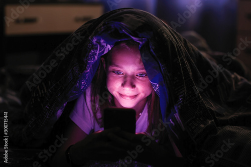 Little cute girl at dark looks at smartphone under a blanket in bed.