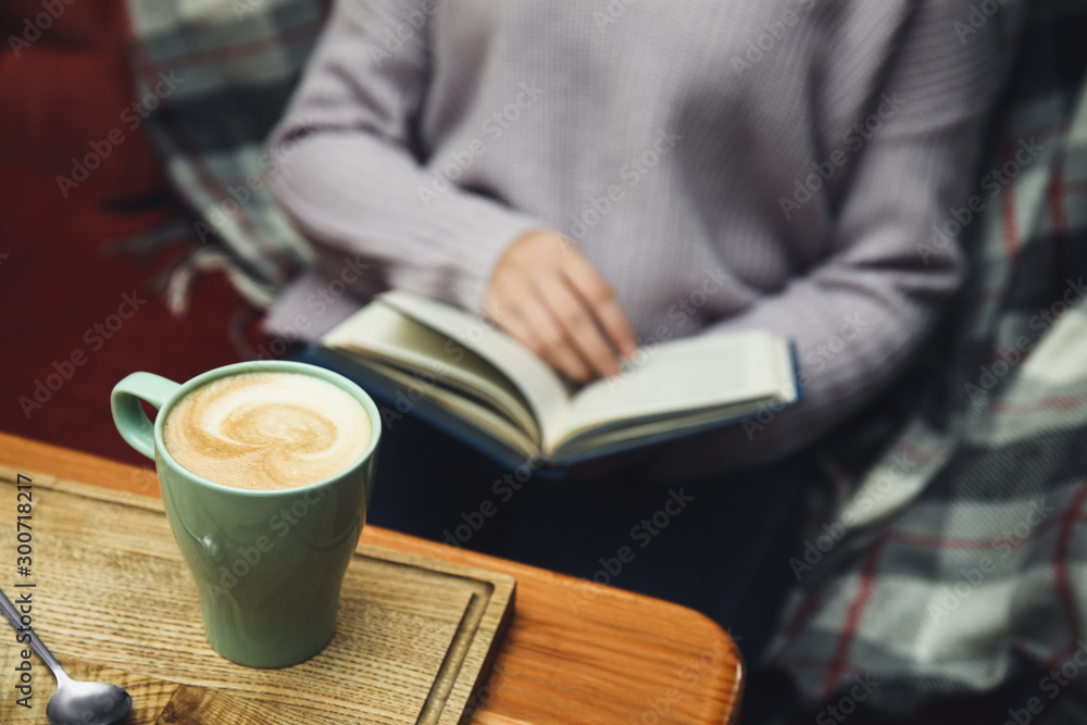 Woman reading book at table, focus on cup of coffee - obrazy, fototapety, plakaty 
