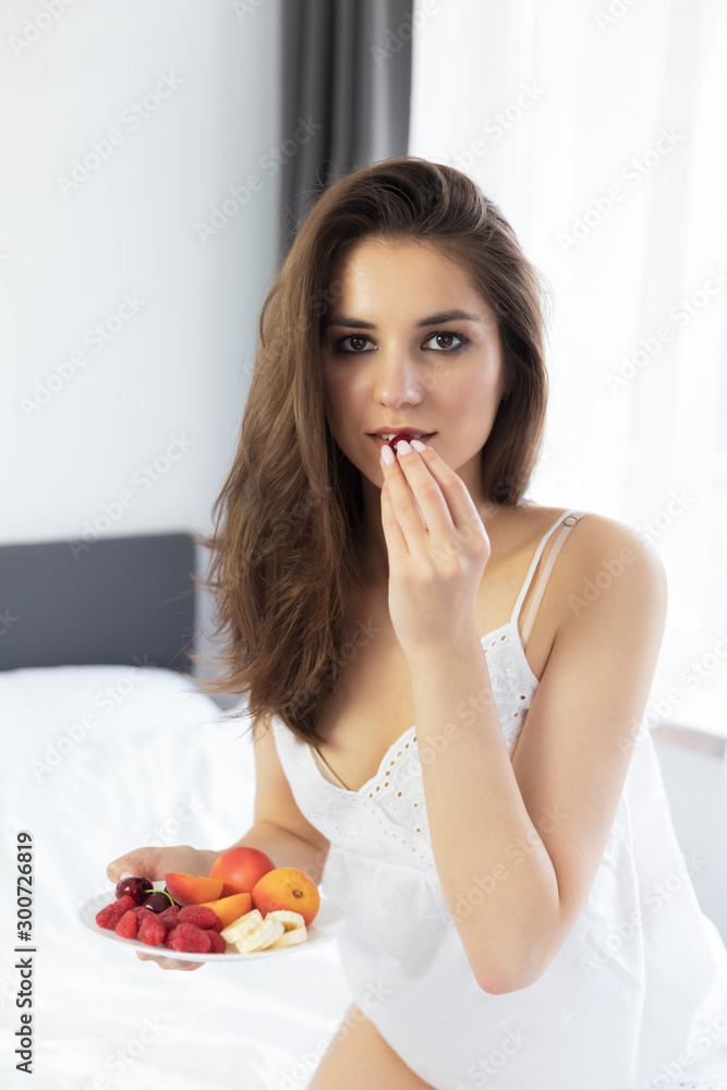 A young attractive girl with long hair is sitting in a white peignoir on the bed.