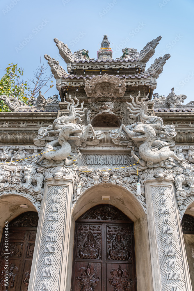 Da Nang, Vietnam - March 10, 2019: Chua An Long Chinese Buddhist Temple. Fisheye closeup on Gray stone Pagoda entrance with brown doors and roof structure under light blue sky and with green foliage.