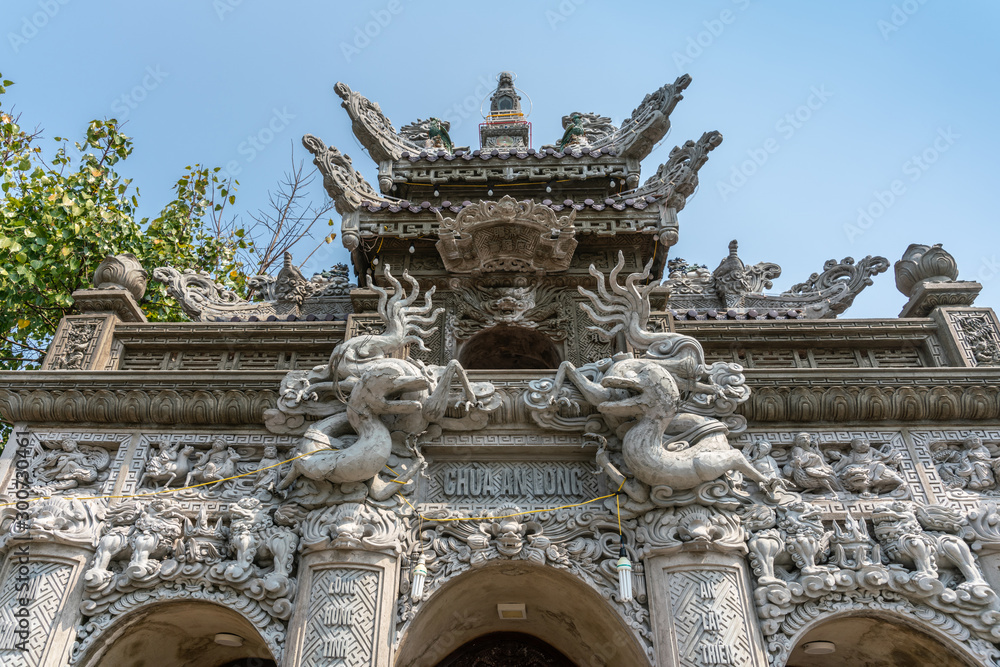 Obraz premium Da Nang, Vietnam - March 10, 2019: Chua An Long Chinese Buddhist Temple. Fisheye on Gray stone Pagoda entrance and roof structure of sanctuary under light blue sky and with green foliage.