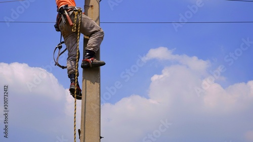 Low section of electrical lineman with tools and equipment is climbing on electric power pole against white cloud and blue sky background