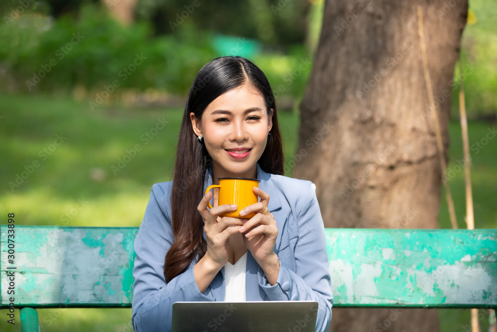 Business women holding coffee cup with laptop sitting on bench in park Business women holding coffee cup with laptop sitting on bench in park