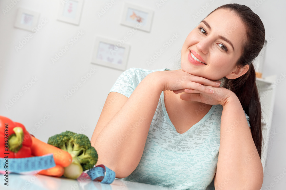 Body Care. Chubby girl sitting at kitchen table with vegetables and tape measure smiling happy close-up Body Care. Chubby girl sitting at kitchen table with vegetables and tape measure smiling happy close-up
