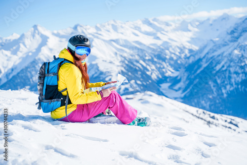 Photo of snowboarder woman in helmet with map in her hands sitting on mountain slope.