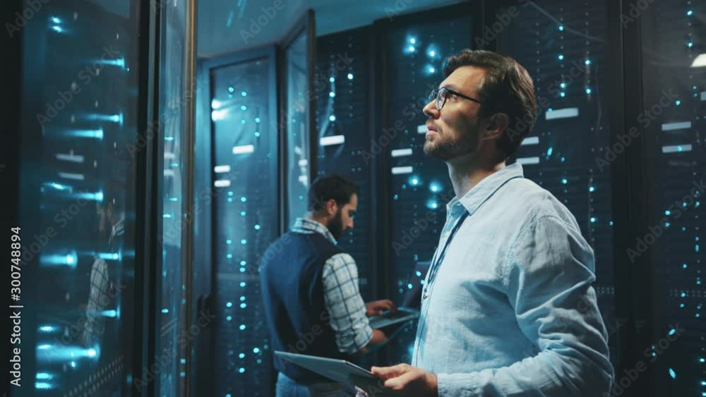 Smart IT engineer working in data center using digital tablet looking into data rack server cabinets running webhosting or maintenance.