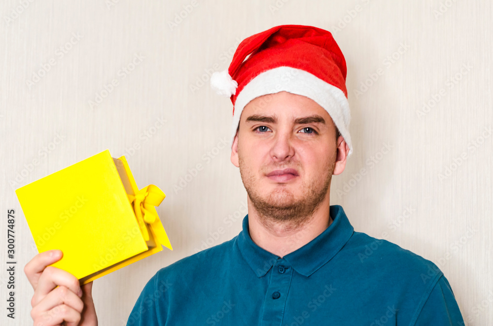 upset, displeased handsome guy in santa hat with disgust holds in his hand a box with gift. young brutal man with negative emotions receives and gives present. spoiled holiday.