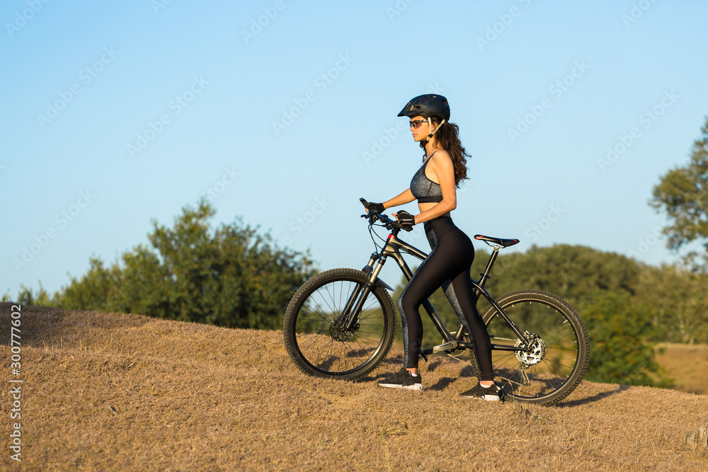 Girl on a mountain bike on offroad, beautiful portrait of a cyclist at sunset, Fitness girl rides a modern carbon fiber mountain bike in sportswear, a helmet, glasses and gloves.	
