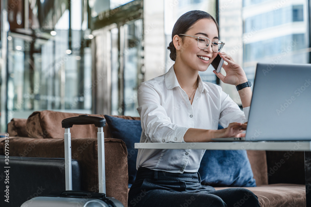 Asian female sitting at airport before trip having telephonic conversation with client and smiling. Asian female sitting at airport before trip having telephonic conversation with client and smiling.