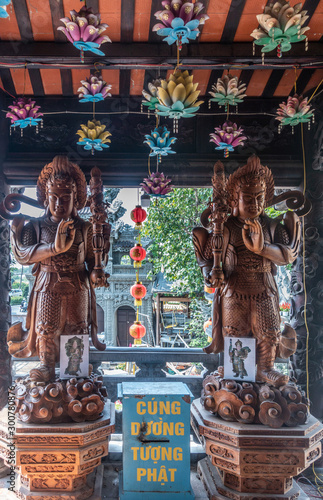 Da Nang, Vietnam - March 10, 2019: Chua An Long Chinese Buddhist Temple. Brown Wooden Heng and Ha guard statues at entrance to sanctuary. Blue donation box and colored lotus on ceiling.