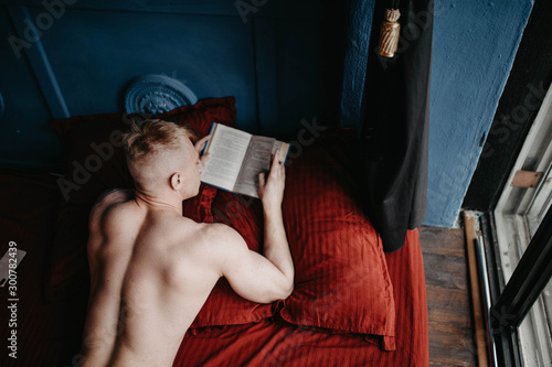 Sexy handsome man with a naked torso is lying in a red bed with a book. photo