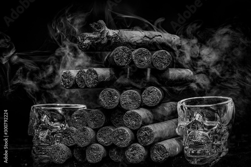 Large stack of hand rolled cuban cigars against dark backround with smoke and human skul shaped shot glasses.