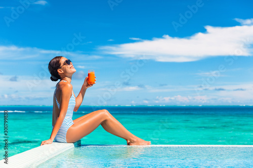 Summer healthy eating woman drinking detox carrot juice for balanced diet weight loss bikini body lifestyle. Happy living girl enjoying sun tan holidays by the swimming pool of tropical hotel.