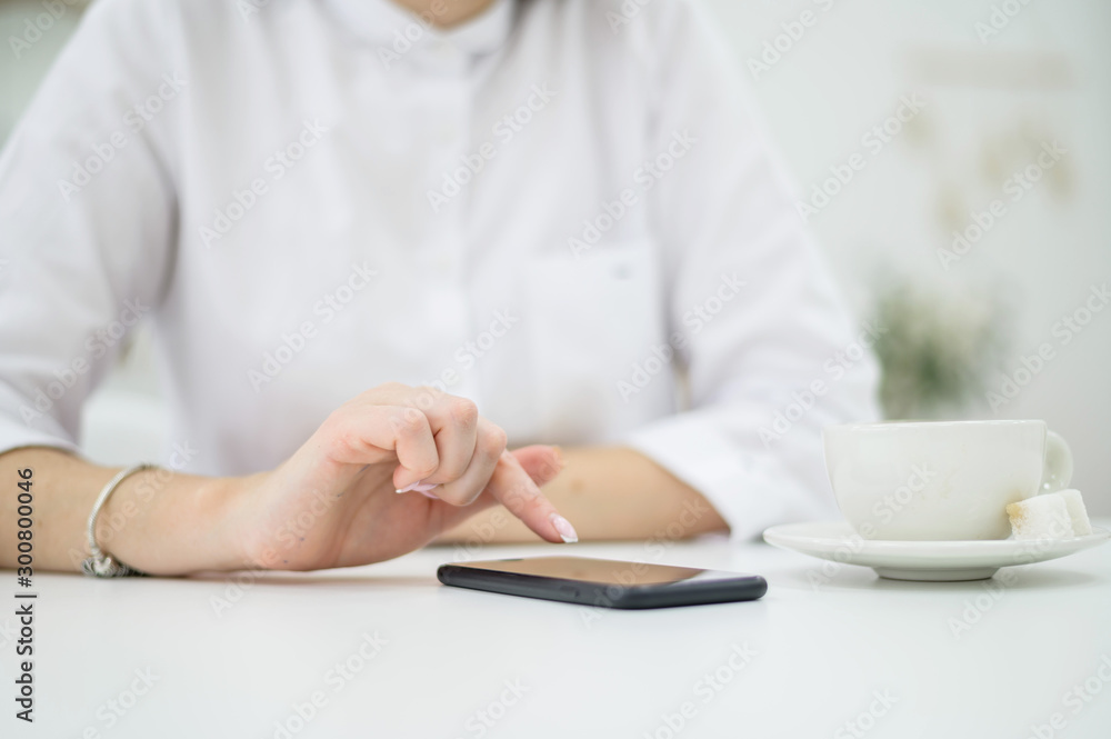 Fototapeta premium A woman in a white shirt with well-groomed hands and fresh manicure is drinking coffee in a cafe. Girl writes a message on a smartphone at lunchtime. Close-up
