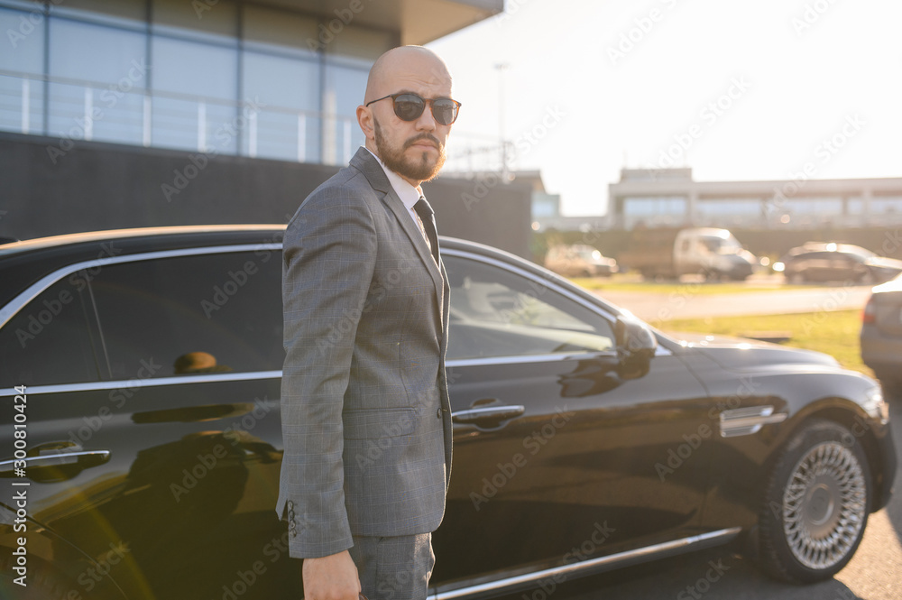 Portrait of a handsome businessman in sunglasses standing near the car outdoors in front of the modern building facade Portrait of a handsome businessman in sunglasses standing near the car outdoors in front of the modern building facade