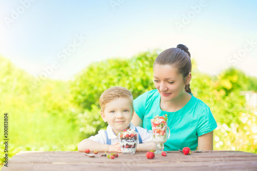Beautiful happy mother and her child boy eating layered dessert in green garden in sunny summer day