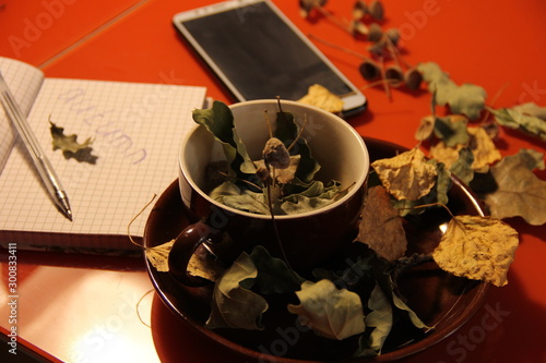 Great autumn background. in a brown mug are dry oak leaves and acorns. Nearby lies a phone - a smartphone and a notepad
