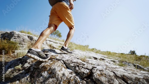 low point wide angle slow motion feet young adult man hiker walking up the rocky slope at summer day against blue sky. slow motion 120 fps back view photo