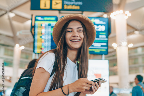 traveler with a backpack and a hat at the airport with a phone smiles cheerfully on the background of the scoreboard
