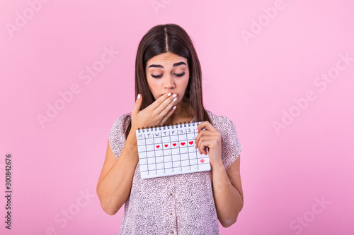 Portrait of a beautiful young girl holding her menstruation calendar and covering hear face isolated over pink background. Period calendar