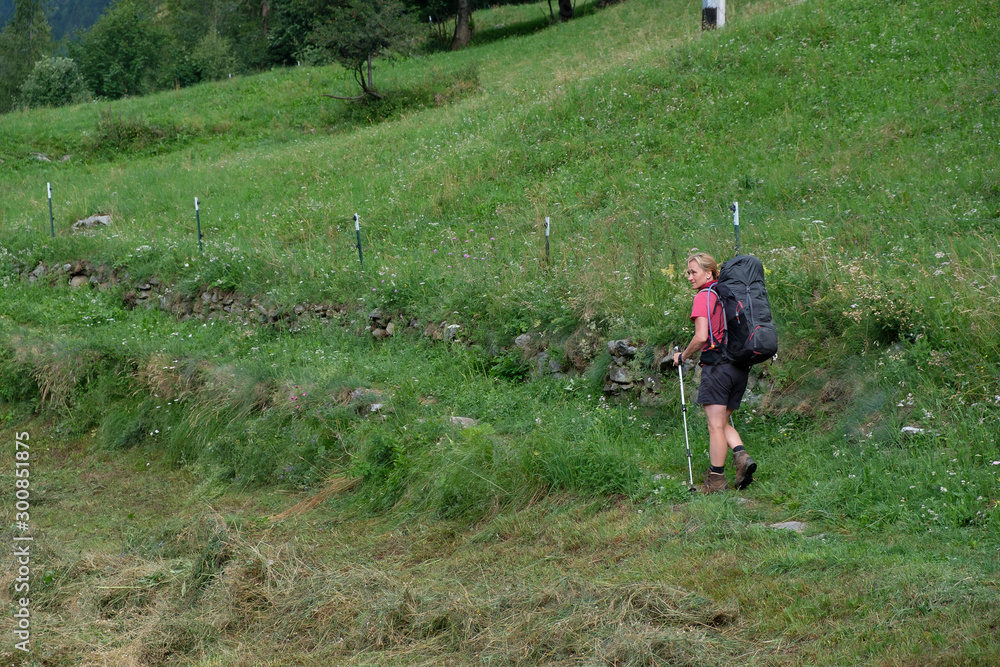 A blond woman with a big backpack wanders through blooming alpine meadows in the Italian Piedmont