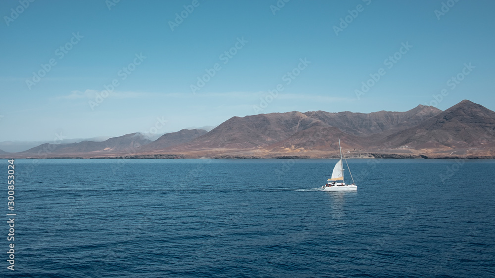 Sailing boat nearby Fuerteventura at its most southern point, at Morro Jable with the display of the solitary cliffs and arid mountains behind, part of the Parque Natural Jandia, Canary Islands, Spain