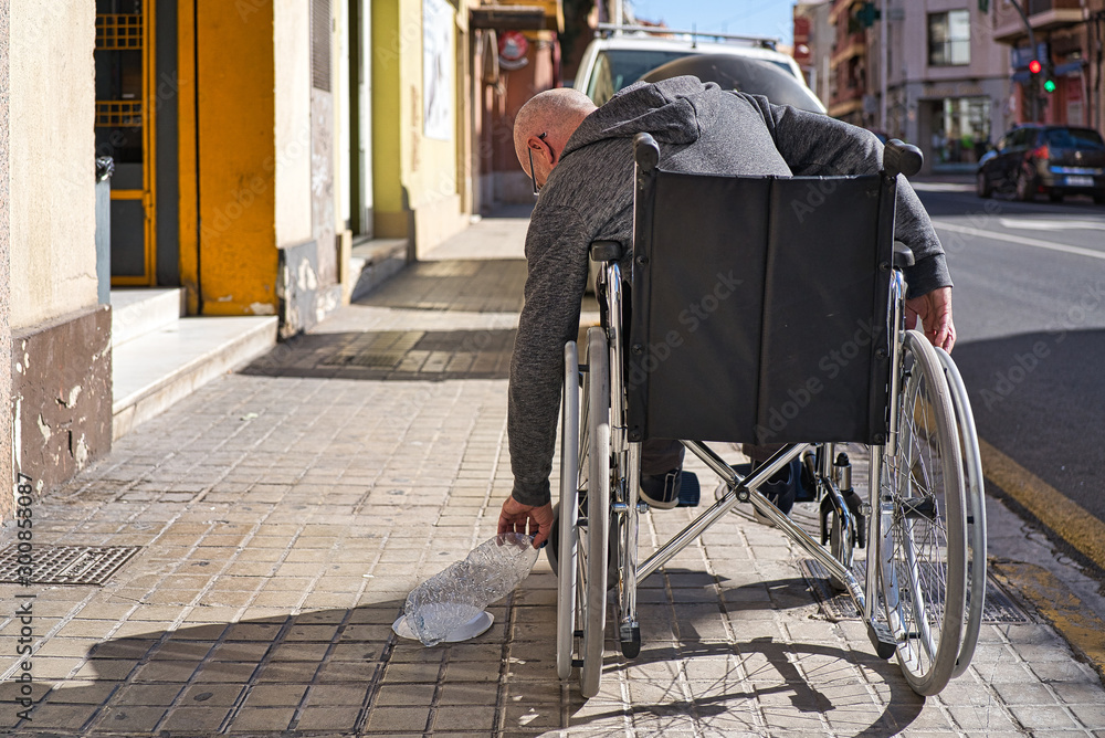 Fototapeta premium Wheelchair disabled person picking up a transparent plastic bottle thrown and abandoned on the floor to throw it into the garbage recycling container and avoid being contaminated. Avoid plastics