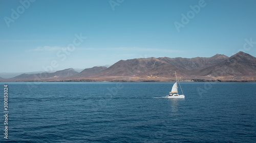 Sailing boat nearby Fuerteventura at its most southern point, at Morro Jable with the display of the solitary cliffs and arid mountains behind, part of the Parque Natural Jandia, Canary Islands, Spain