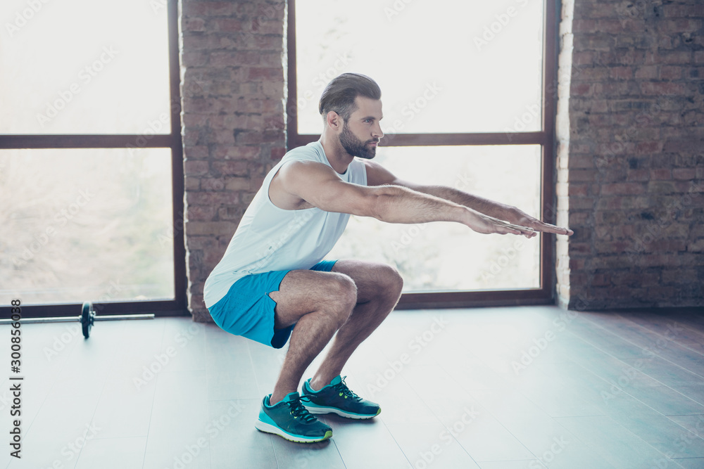Full body profile photo of macho guy doing static squats fat burning process sportswear tank-top shorts sneakers training house home studio near big windows indoors Full body profile photo of macho guy doing static squats fat burning process sportswear tank-top shorts sneakers training house home studio near big windows indoors