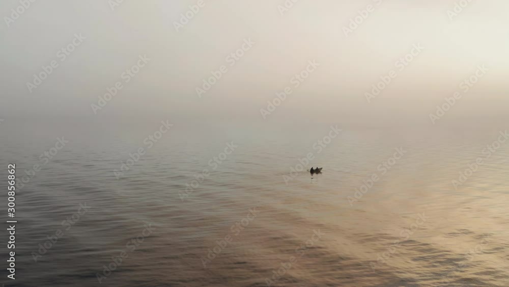 Two fishermen in a rowboat in fog during sunrise on a sea coast - aerial drone shot Two fishermen in a rowboat in fog during sunrise on a sea coast - aerial drone shot