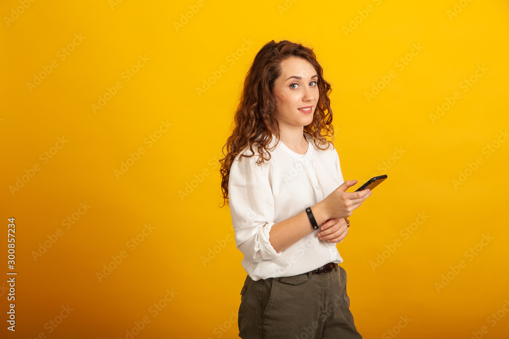 Beautiful young girl with a smartphone in hand looks at the camera and smiles standing isolated on yellow background. Beautiful young girl with a smartphone in hand looks at the camera and smiles standing isolated on yellow background.