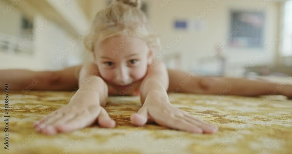Little girl in leotard bending forward and stretching body while doing split on floor in gym