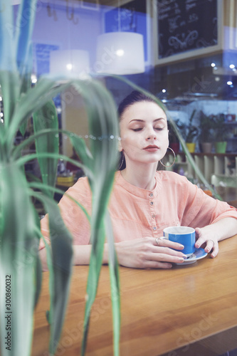 portrait of young woman drinking coffee at table in cafe through the window