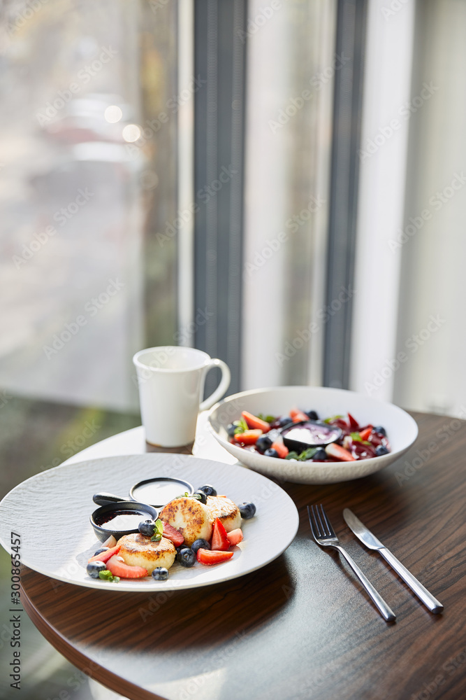 traditional Ukrainian varenyky and syrniki with berries served in white plate with sauce near cutlery and cup on wooden table in restaurant traditional Ukrainian varenyky and syrniki with berries served in white plate with sauce near cutlery and cup on wooden table in restaurant