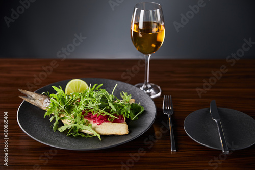tasty restaurant fish steak with lime and arugula on wooden table near cutlery and white wine on black background