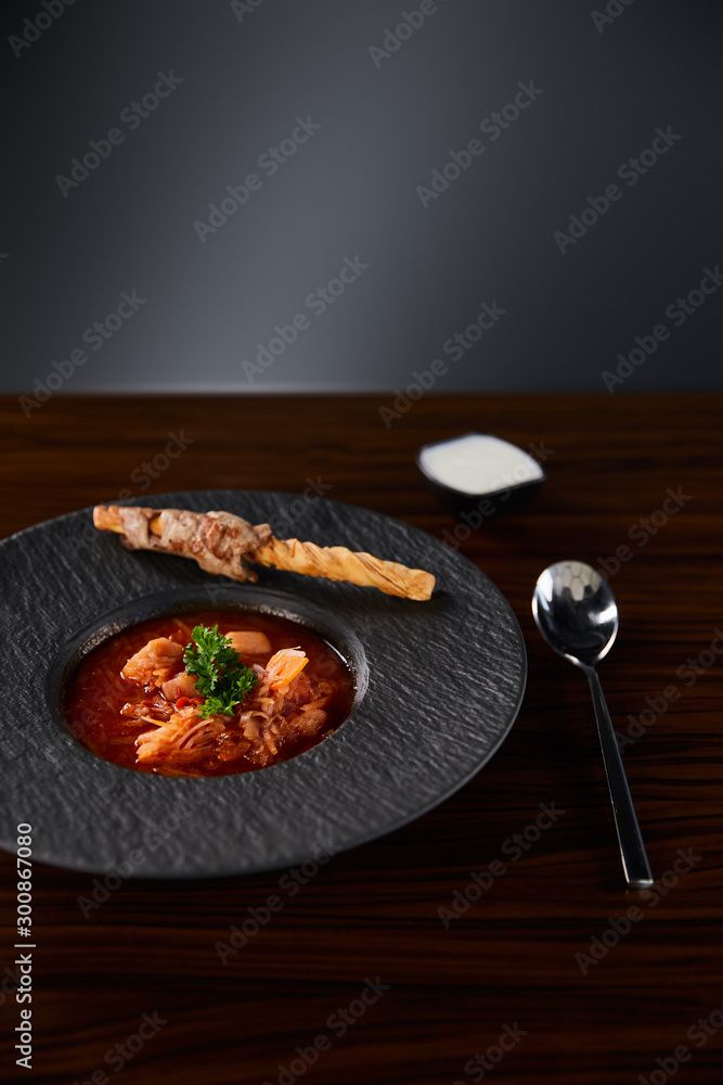 traditional ukrainian borscht garnished with parsley in black plate served with sour cream near spoon at wooden table - obrazy, fototapety, plakaty 