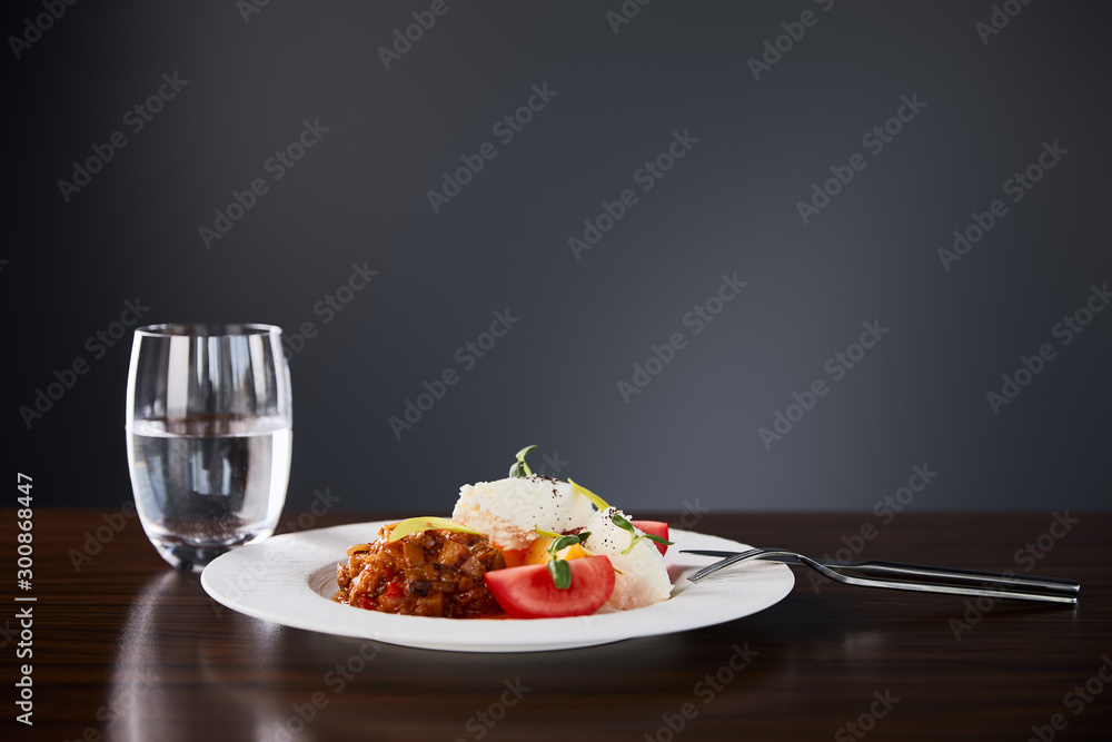 delicious restaurant dish with eggplant caviar and tomatoes served on wooden table with water and cutlery on black background