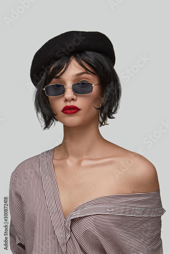 Medium shot of a brunette girl in a striped shirt, black beret and rectangular shaped sunglasses with a goldish metal frame and black lenses. The photo is made on the gray background. photo