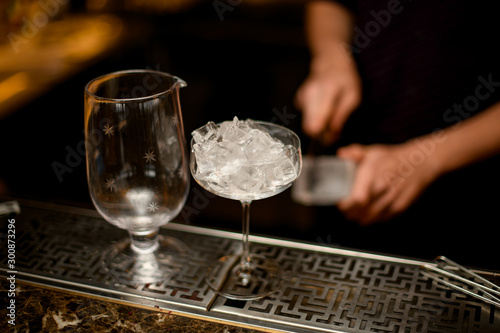 Professional bartender cutting ice with a knife in the foreground of cocktail glasses