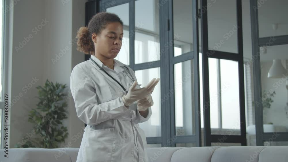 Confident african american female general practitioner putting on sterilized medical gloves to protect and care of patient before medical checkup in domestic interior, looking serious and concentrated Confident african american female general practitioner putting on sterilized medical gloves to protect and care of patient before medical checkup in domestic interior, looking serious and concentrated