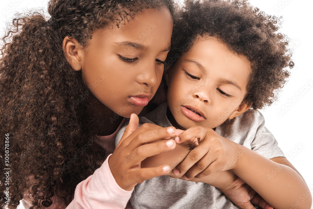 attentive african american sister looking at hand of adorable brother isolated on white attentive african american sister looking at hand of adorable brother isolated on white