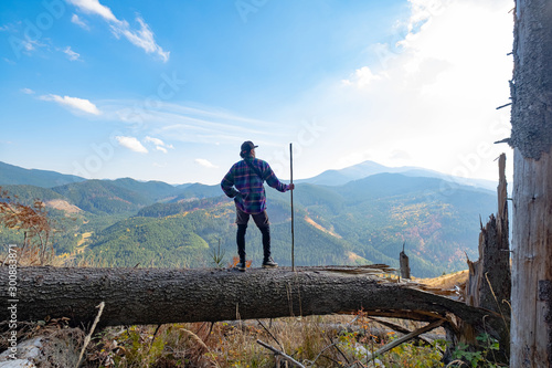 Young male traveler with stick in hands standing on a big log, mountains and sky on background