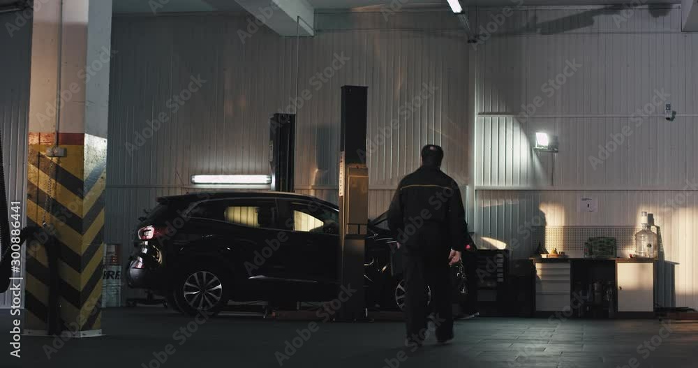 Modern auto service center main mechanic in a uniform going to the other mechanic guy to find the problem of the car Modern auto service center main mechanic in a uniform going to the other mechanic guy to find the problem of the car