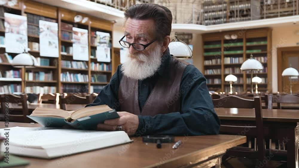 Beautiful portrait of pleasant old bearded man with wrinkled face which putting on his glasses and reading book in library Beautiful portrait of pleasant old bearded man with wrinkled face which putting on his glasses and reading book in library