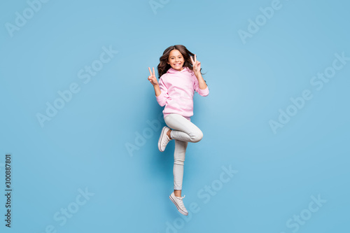 Full length body size photo of cheerful positive preteen showing v-sign smiling pink toothily jumping up isolated pastel blue color background